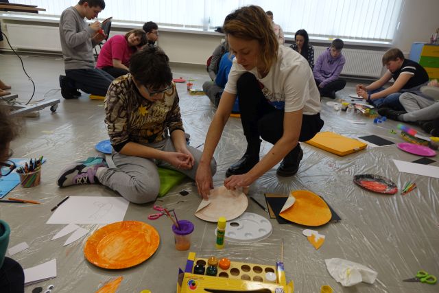 młodzież maluje na papierowych talerzykach, przy nich kuca kobieta