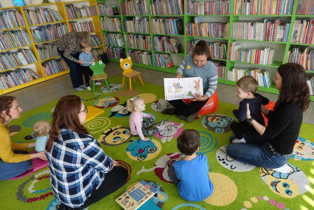 dzieci i osoby dorosłe siedzą na kolorowym dywanie, w tle regały z książkami