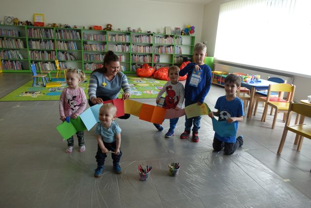 dzieci i kobieta trzymają wykonaną przez siebie długą książkę, w tle regały z książkami