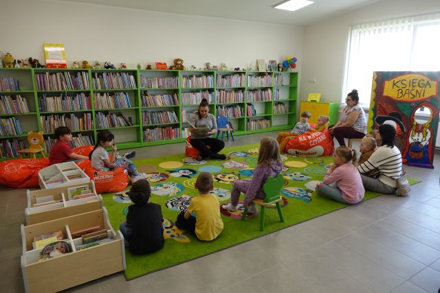 kobieta czyta dzieciom siedzącym na kolorowym dywanie książkę, w tle regały z książkami