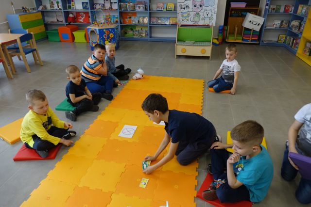 dzieci sterują robotami na pomarańczowej macie, w tle regały