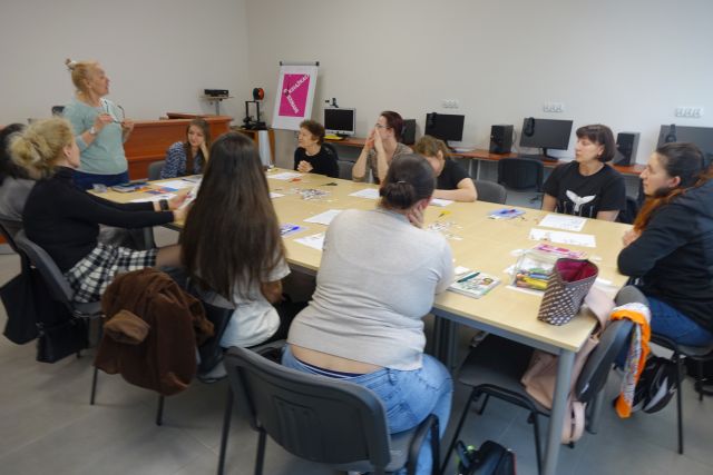 9 kobiet siedzi przy stołach, ma stołach leżą kartki, zeszyty i długopisy, w tle biurka z komputerami