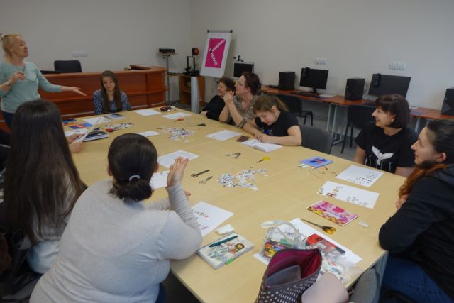 9 kobiet siedzi przy stołach, ma stołach leżą kartki, zeszyty i długopisy, w tle biurka z komputerami