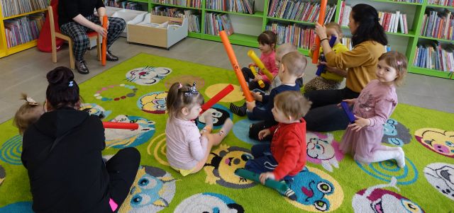 małe dzieci i rodzice bawią się bum bum rurkami siedząc na kolorowym dywanie, w tle regały z książkami