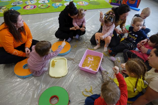 małe dzieci i rodzice siedzą na podłodze i robią korale a makaronu