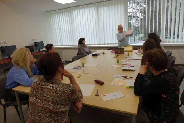 8 kobiet siedzi przy stołach i uczy się polskiego, przed nimi stoi nauczycielka, na stołach kartki