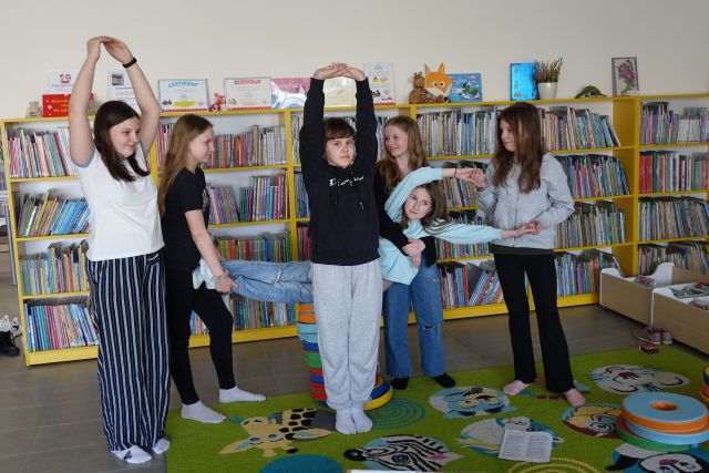 3 dziewczyny trzymają za ręce i nogi dziewczynę, przed nimi stoi dziewczyna z rękoma uniesionymi do góry