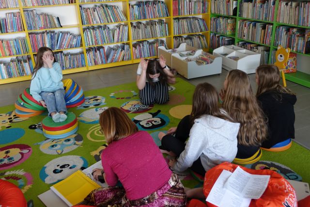 młodzież siedzi pochylona na kolorowym dywanie, w tle regasły z książkami