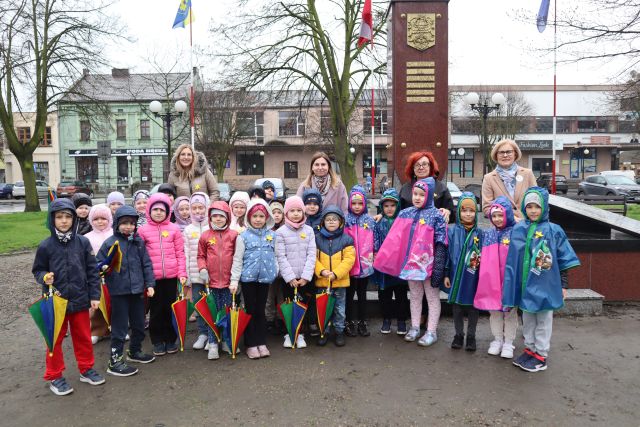 grupa przedszkolaków pozuje do zdjęcia przed pomnikiem, mają przypięte do kurtek żółte żonkile