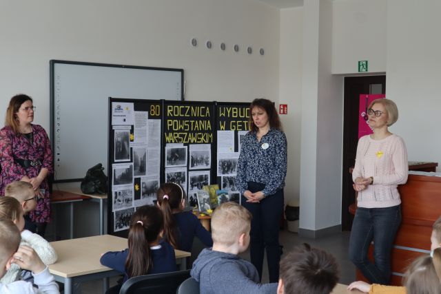 kobieta przemawia do dzieci, w bluzce ma wpiętego żółteko żonkila z papieru, w tle wystawa na temat 80 rocznicy wybuchu powstania w getcie warszawskim