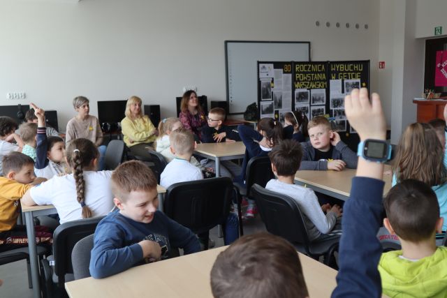 dzieci siedzą przy stolikach, w tle komputery na biurkach