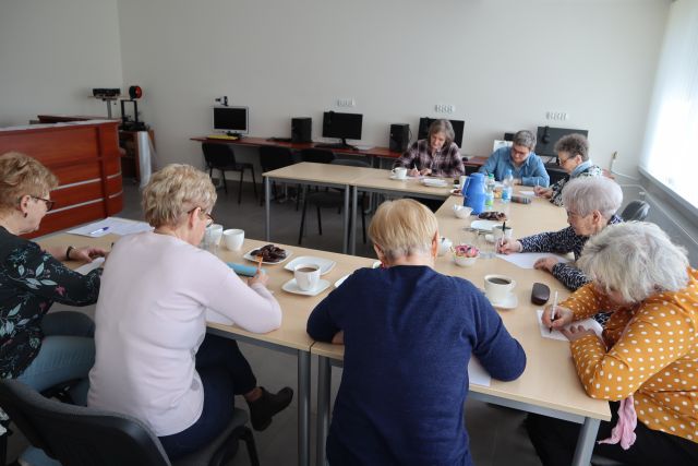 Seniorki siedząc przy stołach rozwiązują łąmigłówki, w tle biurka z kiomputerami