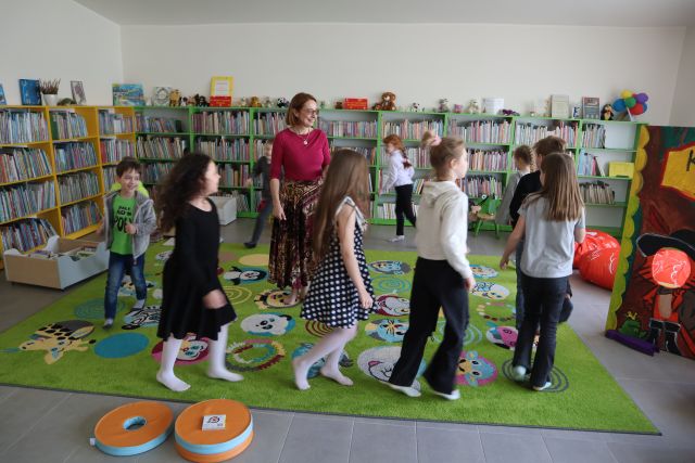 dzieci i prowadząca warsztaty kobieta idą w okręgu po kolorowym dywanie, w tle regały z książkami