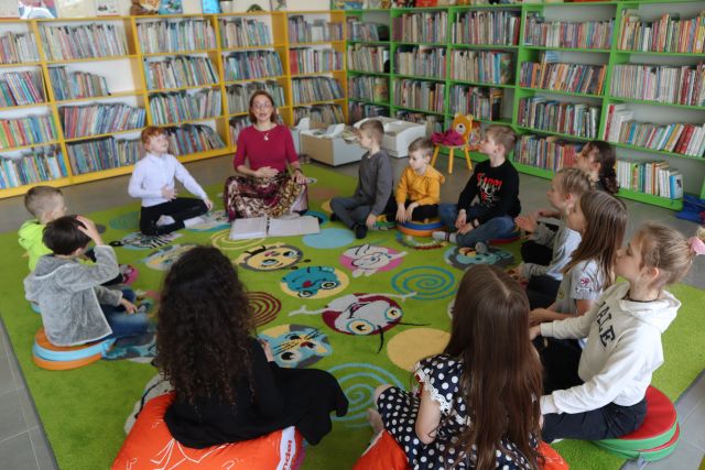 dzieci i kobieta siedzą na kolorowym dywanie, w tle regały z książkami