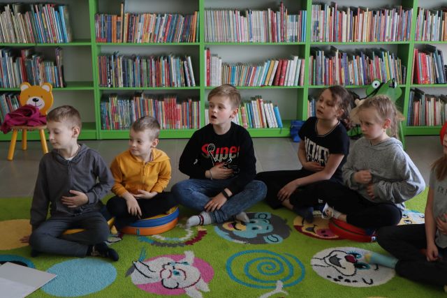 dzieci ćwiczą oddech siedząc na kolorowym dywanie, w tle regały z książkami