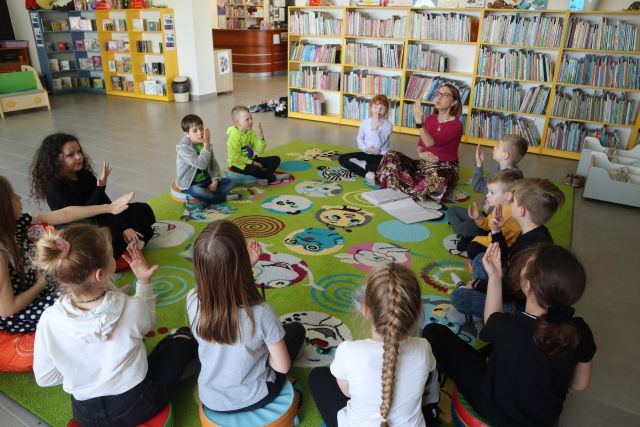 dzieci i kobieta siedzą na kolorowym dywanie i ćwiczą oddech, w tle regały z książkami
