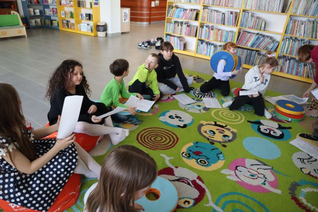 dzieci siedzą na kolorowym dywanie, przed sobą i w rękach mają kartki