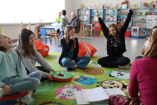 dziewczyny siedzą na kolorowym dywanie, jedna unosi rękę do góry