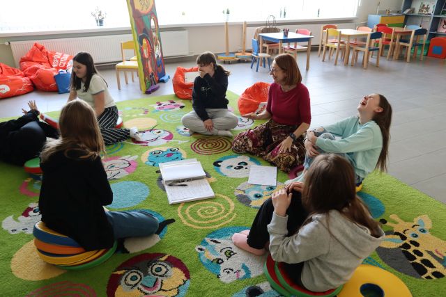 dziewczyny i kobieta prowadząca warsztaty siedzą na kolorowym dywanie, przed nimi zeszyt i kartki