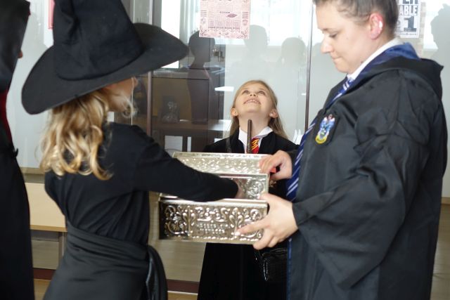 dziewczyna w czarnym kapeluszu losuje ze skrzyni kartkę, przed nią stoi ubrana w czarną pelerynę dziewczynka i kobieta ubrana w czarną pelerynę i czarny kapelusz