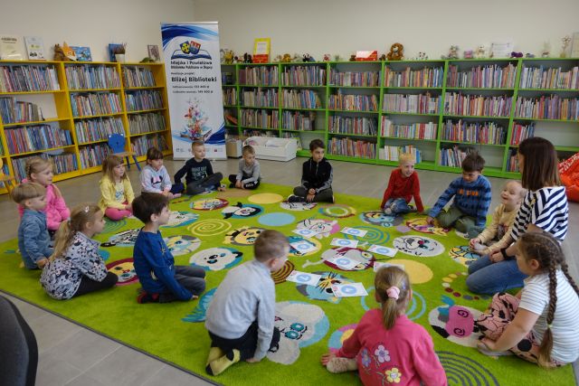dzieci i nauczycielka siedzą na kolorowym dywanie, w tle regały z książkami 