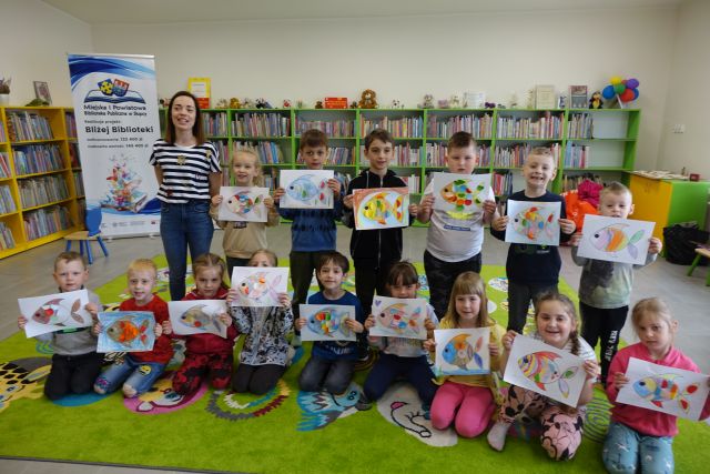 dzieci pozują do zdjęcia z wykonanymi przez siebie kolorowymi rybkami na kartkach, w tle regały z książkami