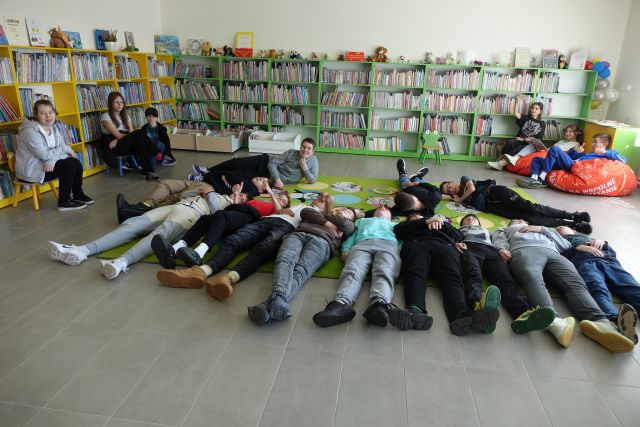 młodzież leży na kolorowym dywanie, w tle regały z książkami