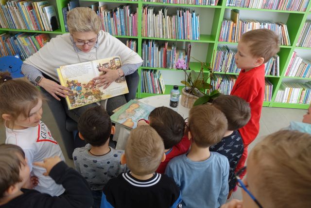 kobieta pokazuje książkę dziciom siedzącym przed nią, za nią regały z książkami