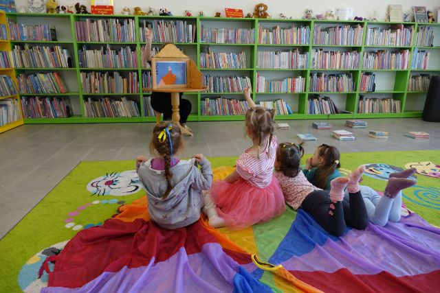 małe dzieci siedzą na kolorowej chuście, przed nimi kobieta czyta w teatrzyku kamishibai