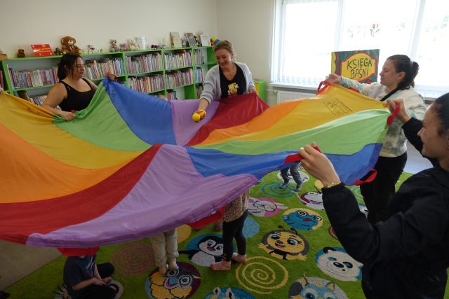 dzieci bawią się pod chustą, a rodzice trzymają chustę i rzucają do niej piłeczki