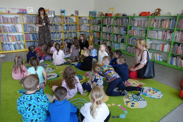 dzieci siedzą na kolorowym dywanie, przed nimi stoi kobieta i opowiada