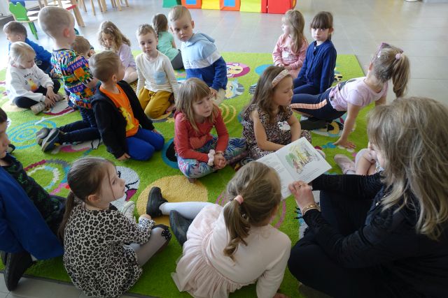 dzieci siedzą na kolorowym dywamnie i słuchają czytającej siedzącej z nimi na dywanie kobiety