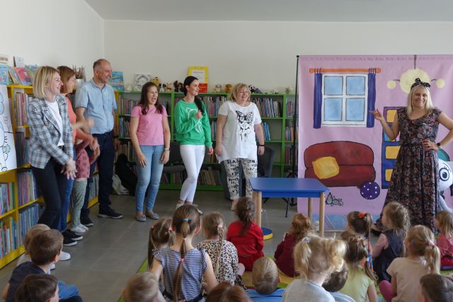 dorośli stoją przed dziećmi siedzącymi na kolorowym dywanie