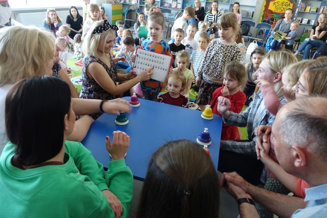 edukatorka pokazuje nuty a dorośli grają na dzwoneczkach, przed nimi na kolorowym dywanie siedzą dzieci