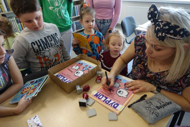 kobieta podpisuje dyplomy, wokół niej stoją dzieci, w tle regały z książkami