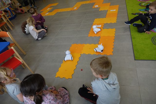 dzieci siedzą na podłodze i sterują przy pomocy tabletów robotami jeżdżącymi po pomarańczowej macie