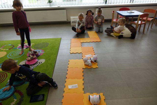 dzieci siedzą na podłodze i sterują przy pomocy tabletów robotami jeżdżącymi po pomarańczowej macie