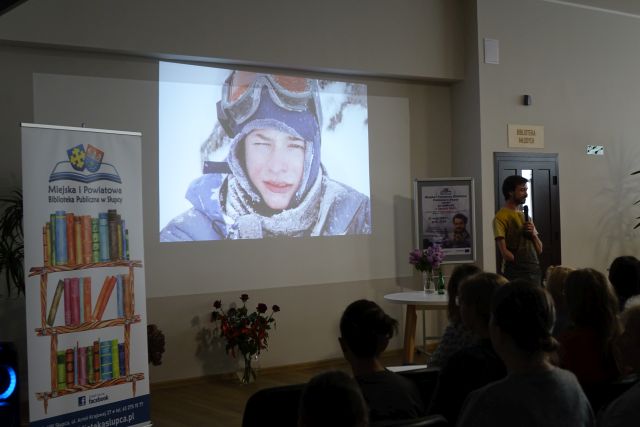 mężczyzna opowiadja stojąc przed publicznością, w tle prezentacja ze zdjęciami z podróży gościa