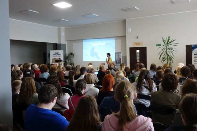 uczestnicy spotkania siedzący na krzesłąch, przed nimi mężczyna opowiadający