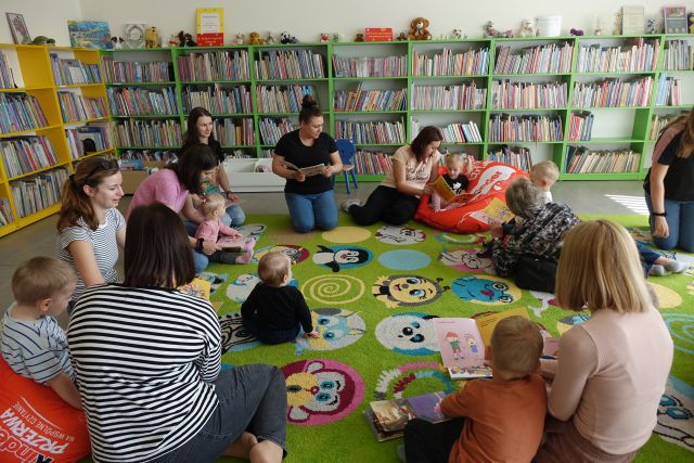 dzieci z rodzicami siedzą na kolorowym dywanie i słuchają opowieści czytanej przez bibliotekarkę, w tle regały z książkami