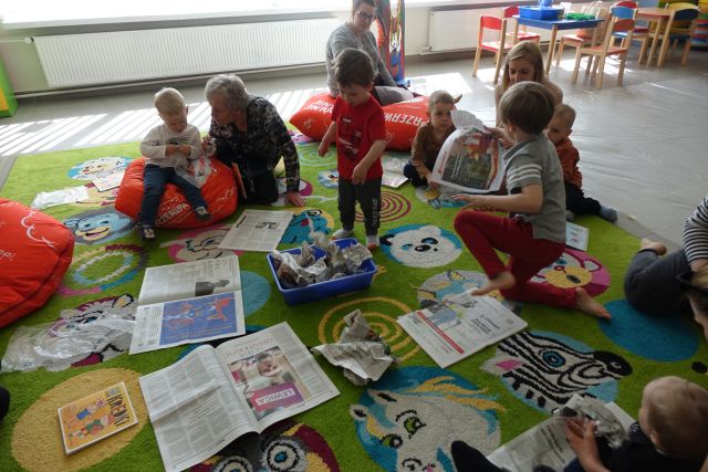 dzieci i rodzice siedzą na kolorowym dywanie, przed nimi rozłożone gazety