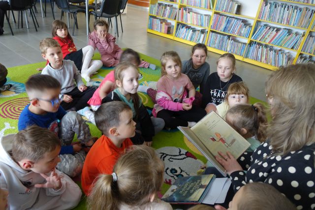 dzieci siedzą na kolorowym dywanie, przed nimi na krzesełku kobirta czyta książkę, w tle regały z książkami