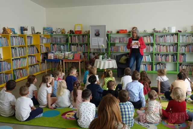 dzieci siedzą na zielonym dywanie przed kobietą stojącą i pokazującą książkę, w tle regały z książkami