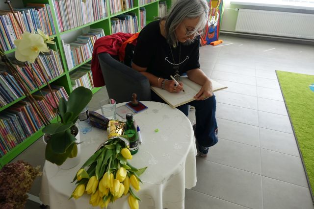 kobieta siedząca na siwym fotelu wpisuje się do kroniki biblioteki, na stoliku przykrytym jasnym obrusem leżą żółte tulipany