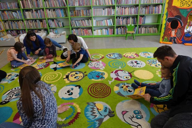 rodzice i dzieci siedzą na kolorowym dywanie i przeglądają książki, w tle regały z książkami