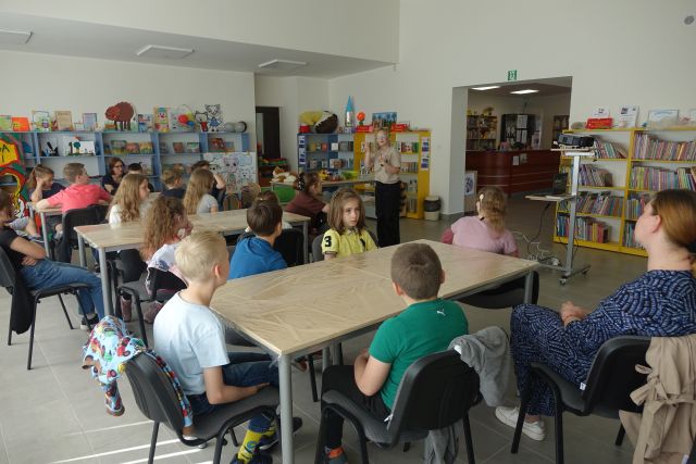 dzieci siedzą przy stolikach, przed nimi stoi i opowiada kobieta