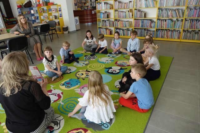 dzieci i kobieta siedzą na kolorowym dywanie, w tle regały z książkami