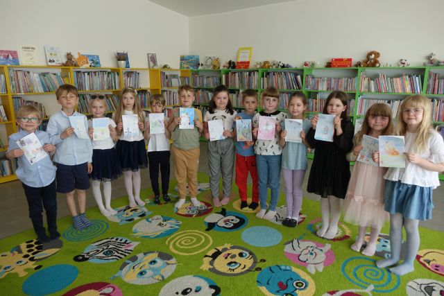dzieci stoją na kolorowym dywanie i prezentują wykonane przez siebie prace, w tle regały z książkami