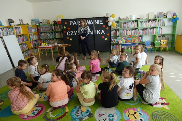 dzieci siedzą na dywanie przed stojącą kobietą, w tle regały z książkami