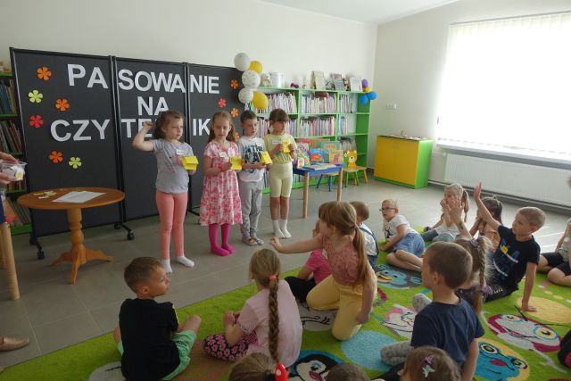 dzieci siedzą na zielonym dywanie, przed nimi stoi trójka dzieci i kobieta